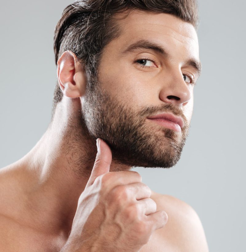 Portrait of a handsome naked bearded man examining his face isolated over white background